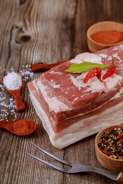 Primo piano del pezzo di pancia di maiale crudo su una superficie di legno con spezie ed erbe aromatiche