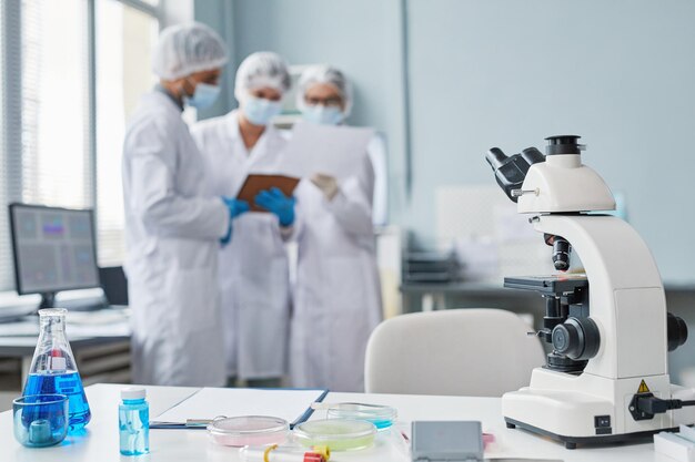 Primo piano del posto di lavoro dello scienziato con microscopio e vetreria di laboratorio con liquido sul tavolo con persone sullo sfondo