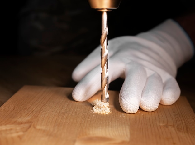 Primo piano del processo di perforazione. Strumento di perforazione in acciaio di riparazione del lavoro manuale che fa foro in tavola di legno e mani maschili in guanti.