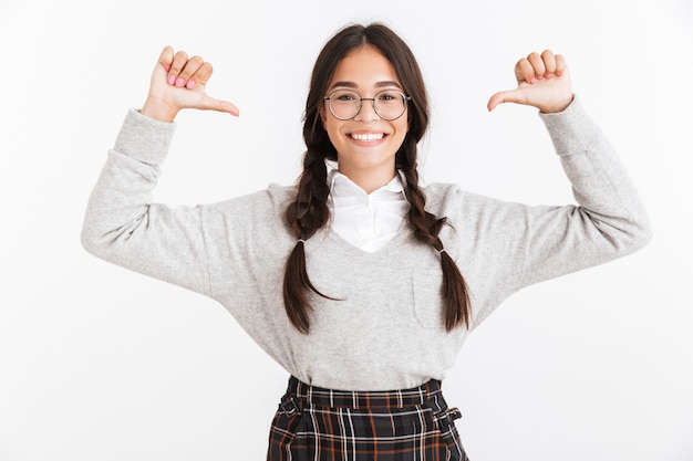 Primo piano del ritratto dell'adolescente adorabile che indossa gli occhiali che sorride e che indica le dita a se stessa isolata sopra la parete bianca