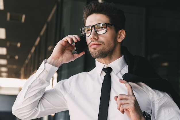 Primo piano del ritratto di giovane uomo d'affari vestito in abito formale in piedi fuori dall'edificio di vetro con la giacca sulla spalla e parlando al telefono cellulare