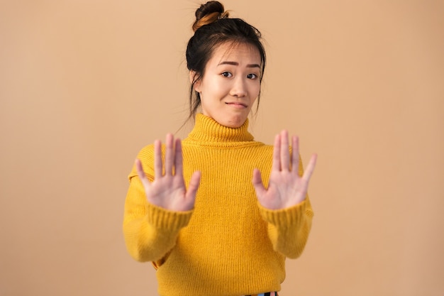 Primo piano del ritratto di una donna cinese seria che indossa un maglione facendo un gesto di arresto con le mani isolate sul muro beige in studio