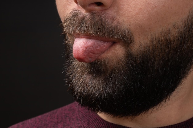 Primo piano del ritratto ritagliato di giovane uomo barbuto che mostra la lingua fuori isolato su sfondo nero emo...