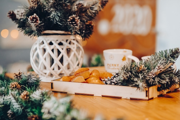 Primo piano del vassoio in legno con gustosi cupcakes fatti in casa e rami di abete sul tavolo con vaso di rami con pigne. Bokeh.