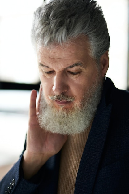 Primo piano del viso di un uomo di mezza età dai capelli grigi elegante e pensieroso che guarda in basso toccandosi la barba