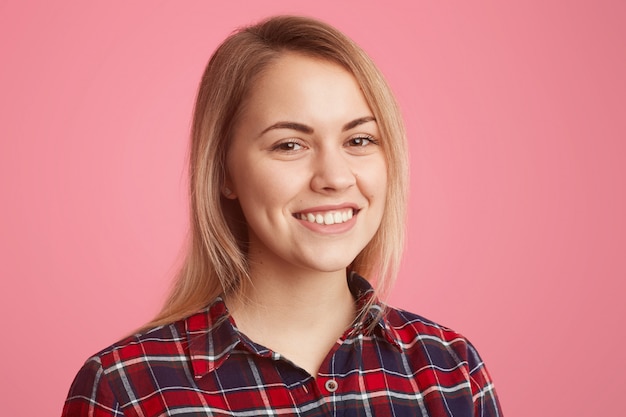 Primo piano del volto di una giovane donna europea bionda dall'aspetto piacevole con un sorriso a trentadue denti, trucco minimo, indossa una camicia a scacchi, gode di una buona atmosfera, essendo di buon umore, isolata sul rosa