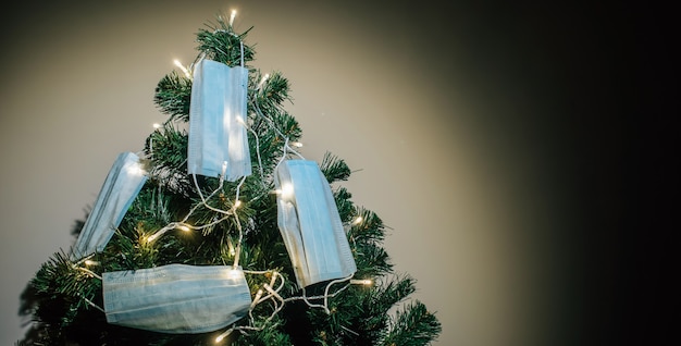 Primo piano dell'albero di Natale illuminato con ghirlande, decorato con maschere mediche protettive in una stanza buia. ¡oncept di celebrare il nuovo anno 2021 durante la pandemia di Covid-19. Copia spazio.