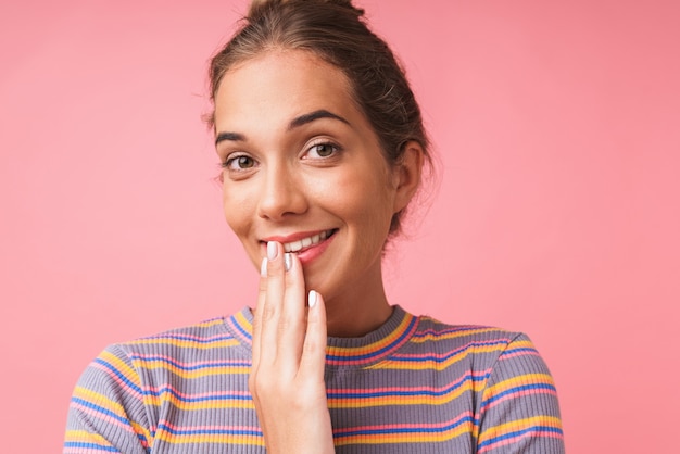 Primo piano dell'immagine di una bella donna felice vestita con abiti colorati che sorride e si copre la bocca isolata sul muro rosa