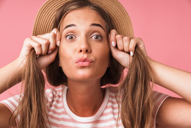 Primo piano dell'immagine di una donna allegra e carina che indossa un cappello di paglia che si prende in giro con i suoi capelli e fa una smorfia isolata sul muro rosa