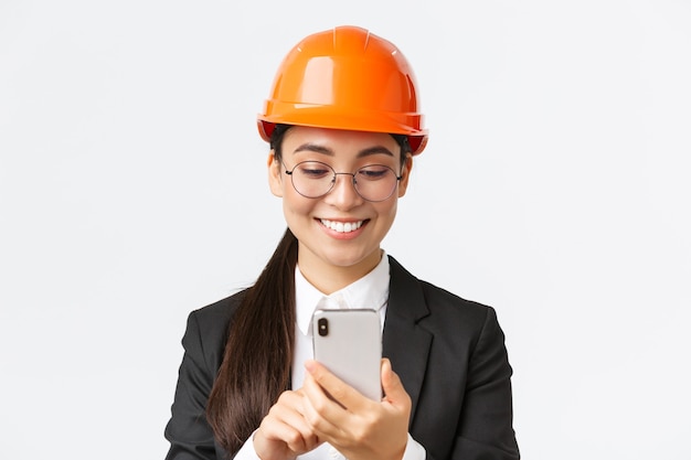 Primo piano dell'ingegnere industriale femminile asiatico professionale sorridente, indossando il casco di sicurezza e la tuta da lavoro, utilizzando lo smartphone, guardando soddisfatto lo schermo del telefono cellulare, in piedi sfondo bianco