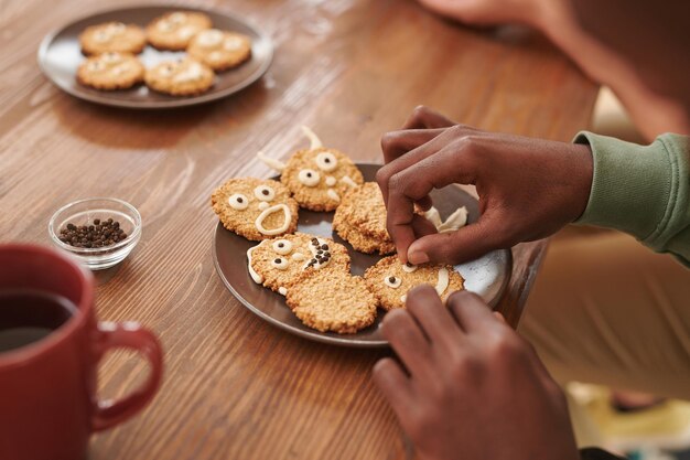 Primo piano dell'uomo afroamericano seduto al tavolo di legno e aggiungendo un design divertente sui biscotti di zucchero mentre