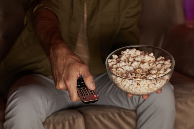 Primo piano dell'uomo che cambia i canali in TV mentre guarda la TV e mangia popcorn