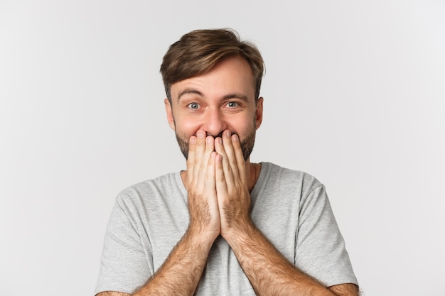 Primo piano dell'uomo eccitato e sorpreso, coprire la bocca e ridere, in piedi sopra il bianco.