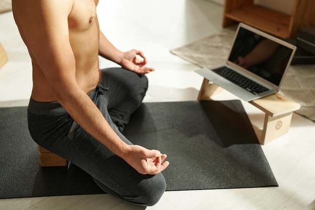 Primo piano dell'uomo senza camicia seduto nella posizione del loto sul tappetino per esercizi davanti al computer portatile e facendo yoga