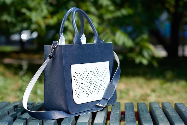 Primo piano della borsa da donna abbastanza alla moda, realizzata in due colori: blu e bianco. Sta sulla panchina del parco. Include cintura lunga e tasca goffrata. La foto è stata fatta su uno sfondo bianco.