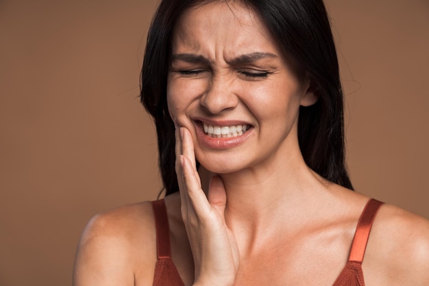 Primo piano della donna bruna che preme la sua guancia con un'espressione dolorosa perché ha un terribile mal di denti, isolata su uno sfondo di colore beige