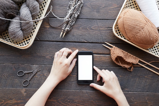 Primo piano della donna che utilizza il suo telefono cellulare seduto al tavolo con gomitoli di lana che guarda lezione di maglieria online