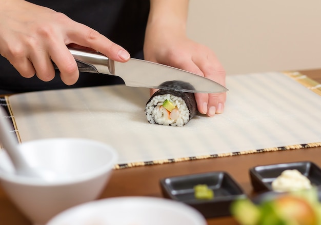 Primo piano della donna chef che taglia rotoli di sushi giapponesi con riso, avocado e gamberetti su un foglio di alga nori