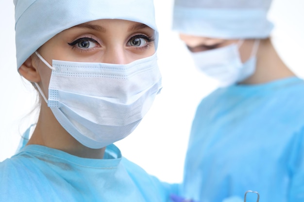 Primo piano della donna chirurgo che guarda la fotocamera mentre i colleghi si esibiscono in background nella sala operatoria, isolata. Concetto di chirurgia e medicina
