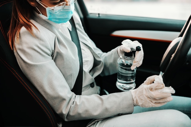 Primo piano della donna con maschera facciale con guanti di gomma sul volante di spruzzatura con alcool e disinfettare la sua auto durante l'epidemia di virus corona.