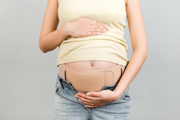 Primo piano della donna incinta che indossa corsetto gravidanza contro il dolore alla schiena in superficie grigia con spazio di copia