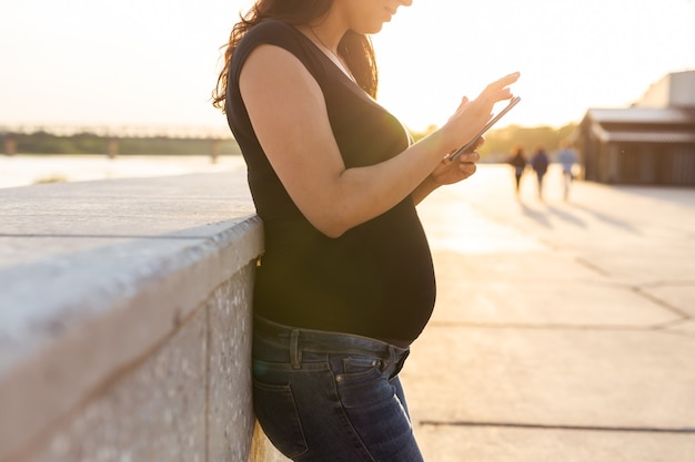 Primo piano della donna incinta con lo smartphone al parco. Gravidanza, tecnologia e concetto di comunicazione.