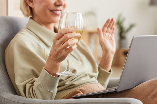 Primo piano della donna sorridente che beve vino e che parla in linea con la sua amica utilizzando il computer portatile a casa