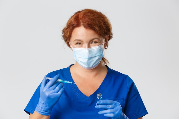 Primo piano della dottoressa gentile sorridente in maschera e guanti di gomma che tengono il vaccino da virus e siringa, andando a fare colpo, iniezione.