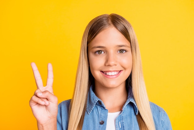 Primo piano della foto del primo piano della scolaretta bionda sorridente che mostra la dimostrazione del segno V con due dita isolate su uno sfondo di colore giallo brillante