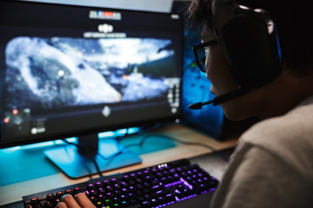 Primo piano della foto del ragazzo asiatico del giocatore che gioca ai videogiochi online sul computer in camera oscura, indossa le cuffie con microfono e utilizza la tastiera colorata retroilluminata