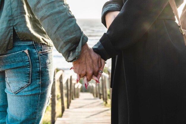 Primo piano della giovane coppia che si tiene per mano camminando giù sulla scala di legno verso la spiaggia Coppia hipster che si tiene per mano innamorata godendosi una serata romantica in spiaggia Mano femminile con belle unghie lunghe