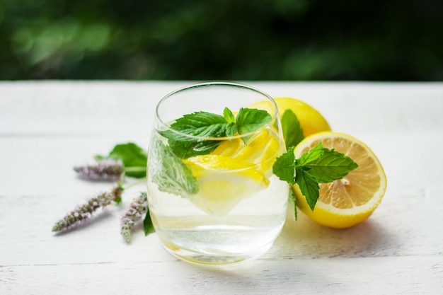 Primo piano della limonata fatta in casa con menta al limone e cubetti di ghiaccio sul tavolo di legno bianco con limoni e fiori di menta