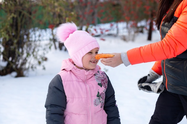 Primo piano della mano adulta femminile che mette la carota alla faccia della figlia come naso sul cortile pieno di neve la sera che cammina con gli alberi e la recinzione di ferro sullo sfondo I genitori trascorrono del tempo con i bambini