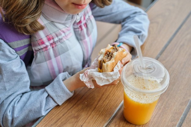 Primo piano della mano del bambino che mangia hamburger e che beve il succo di arancia