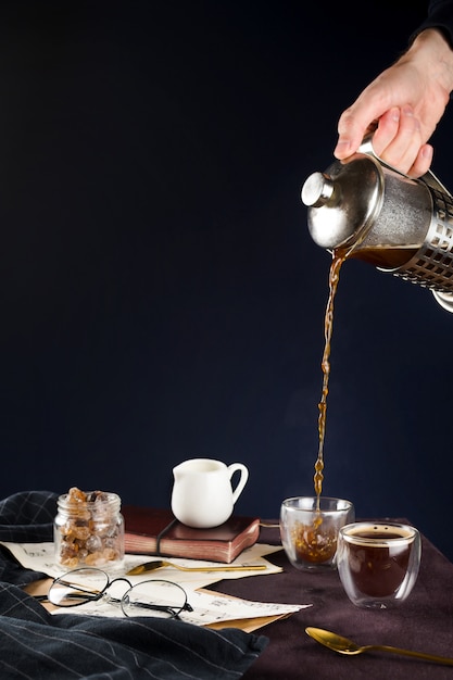 Primo piano della mano dell'uomo versando il caffè nero dalla stampa francese in una tazza su un tavolo di legno con bicchieri, porta latte, zucchero di canna e taccuino, messa a fuoco selettiva