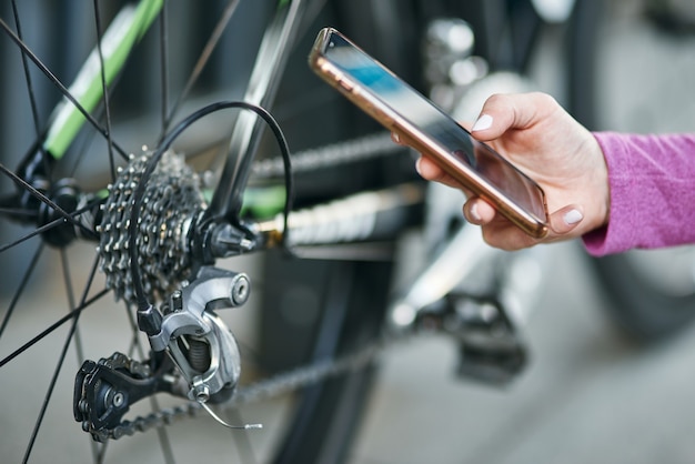 Primo piano della mano della ciclista che tiene lo smartphone mentre controlla i meccanismi della bicicletta, la ruota dentata e la catena su una mountain bike all'aperto durante il giorno. Sicurezza, sport, concetto di tecnologia