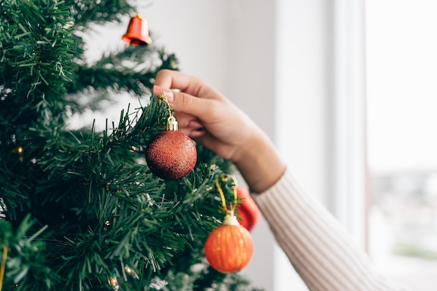 Primo piano della mano della donna che tocca e decora una pallina rossa sull'albero di Natale nel soggiorno di casa Godendo e celebrando le vacanze di Natale