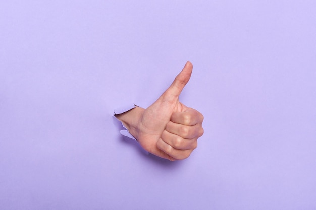 Primo piano della mano della persona con il pollice in su attraverso il foro nella parete di carta viola che dimostra approvazione come gesto di soddisfazione del lavoro svolto Banner per le vendite