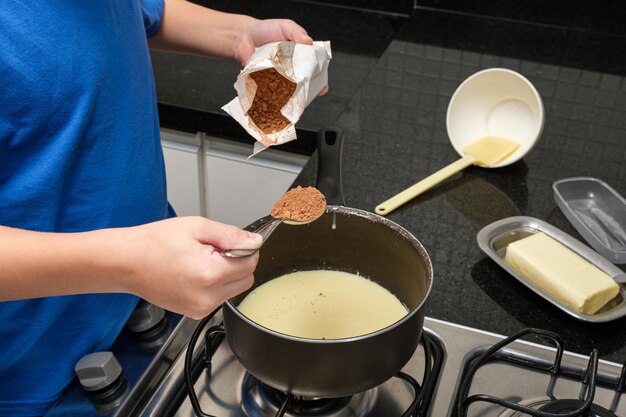 Primo piano della mano di un bambino che versa un cucchiaio di cacao in polvere nel latte condensato per fare il brigadeiro brigadiere