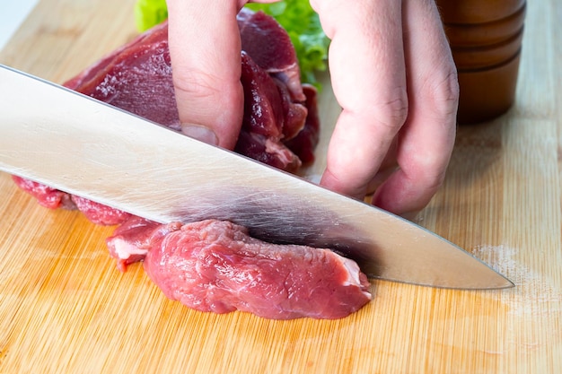 Primo piano della mano di un cuoco unico che taglia la carne di manzo cruda con il coltello in una cucina