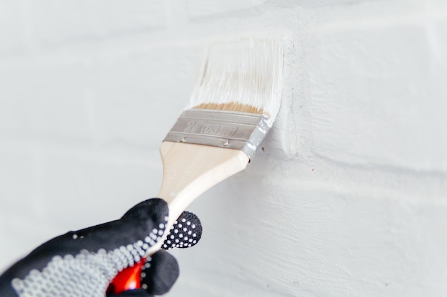 Primo piano della mano di un lavoratore con un pennello che dipinge un muro di mattoni con vernice bianca