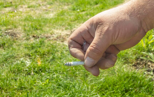 Primo piano della mano di un uomo che fuma una sigaretta su uno sfondo di erba all'aperto per smettere di fumare spazio concettuale per il testo a sinistra