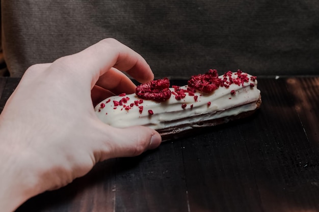 Primo piano della mano di una donna che raggiunge un dolce francese tradizionale eclair con cioccolato bianco e lamponi su sfondo scuro