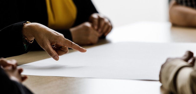 Primo piano della mano di una donna d'affari irriconoscibile non identificata che punta su carta bianca sul tavolo della riunione nella sala conferenze mentre altri discutono della pianificazione della strategia aziendale come lavoro di squadra in compagnia.