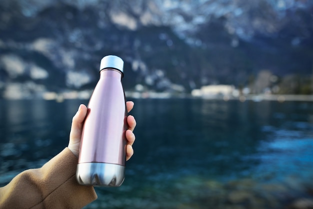 Primo piano della mano femminile che tiene la bottiglia riutilizzabile, in acciaio termo lucido per acqua, sullo sfondo dell'acqua limpida di un lago.