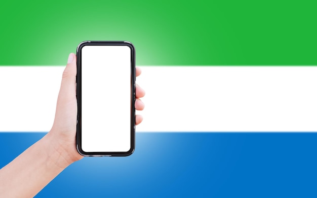 Primo piano della mano maschile che tiene lo smartphone con vuoto sullo schermo sullo sfondo della bandiera sfocata della Sierra Leone