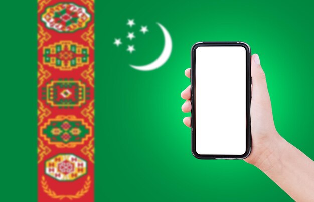 Primo piano della mano maschile che tiene smartphone con schermo vuoto sullo sfondo della bandiera sfocata del Turkmenistan