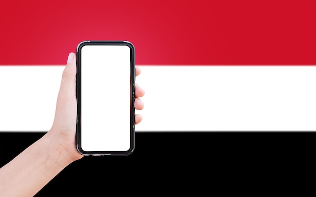 Primo piano della mano maschile che tiene smartphone con schermo vuoto sullo sfondo della bandiera sfocata dello Yemen