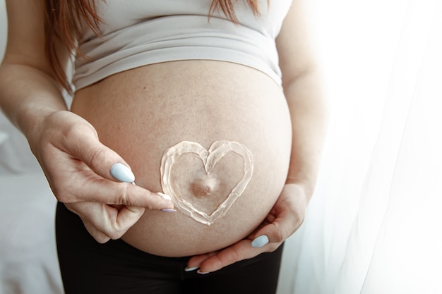 Primo piano della pancia nuda di una futura mamma negli ultimi mesi di gravidanza con un cuore dipinto di crema.