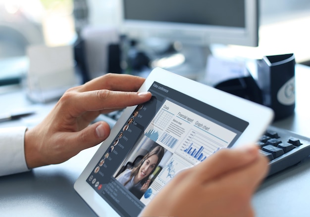 Primo piano della persona in videoconferenza con i colleghi su tablet digitale, analizzando le statistiche finanziarie visualizzate sullo schermo del laptop.